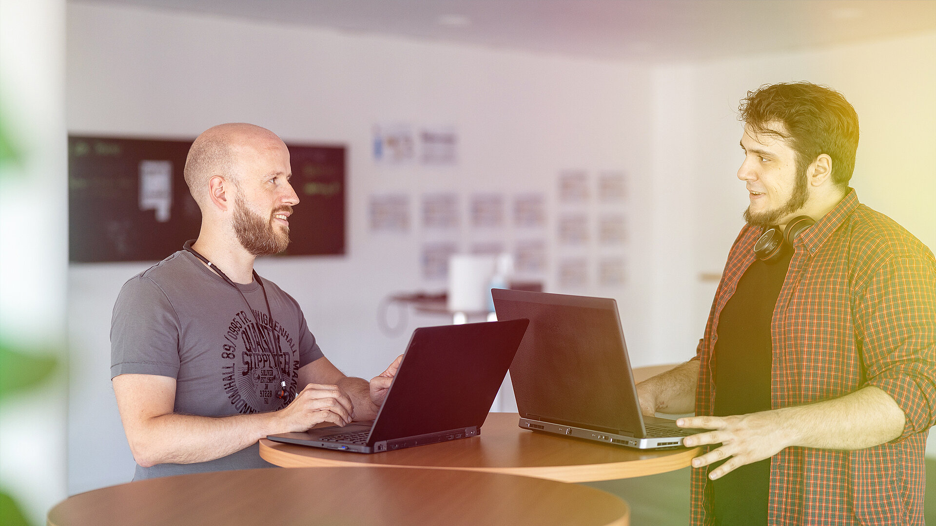 Zwei Männer sitzen an einem Tisch mit Laptops und unterhalten sich.