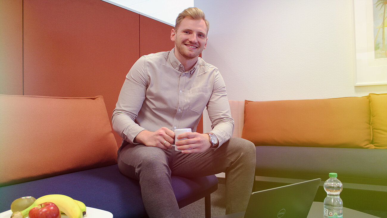 Ein lächelnder Mann sitzt auf einem Sofa mit einem Laptop und einer Tasse Kaffee.