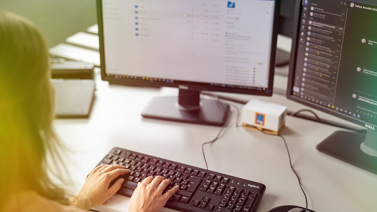 Eine Frau tippt auf einer Tastatur vor zwei Computermonitoren.