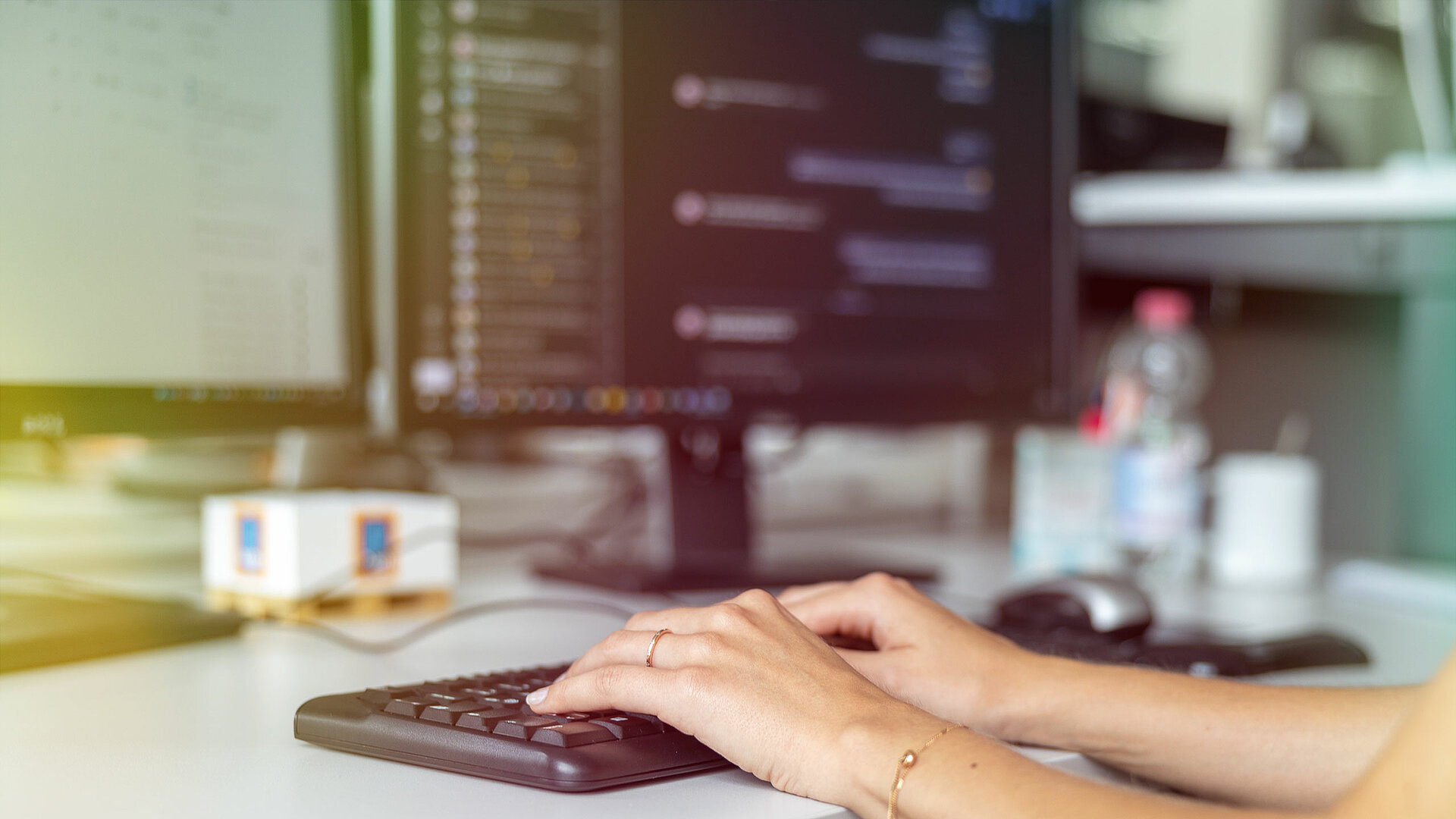 Eine Person tippt auf einer Tastatur vor zwei Computermonitoren.